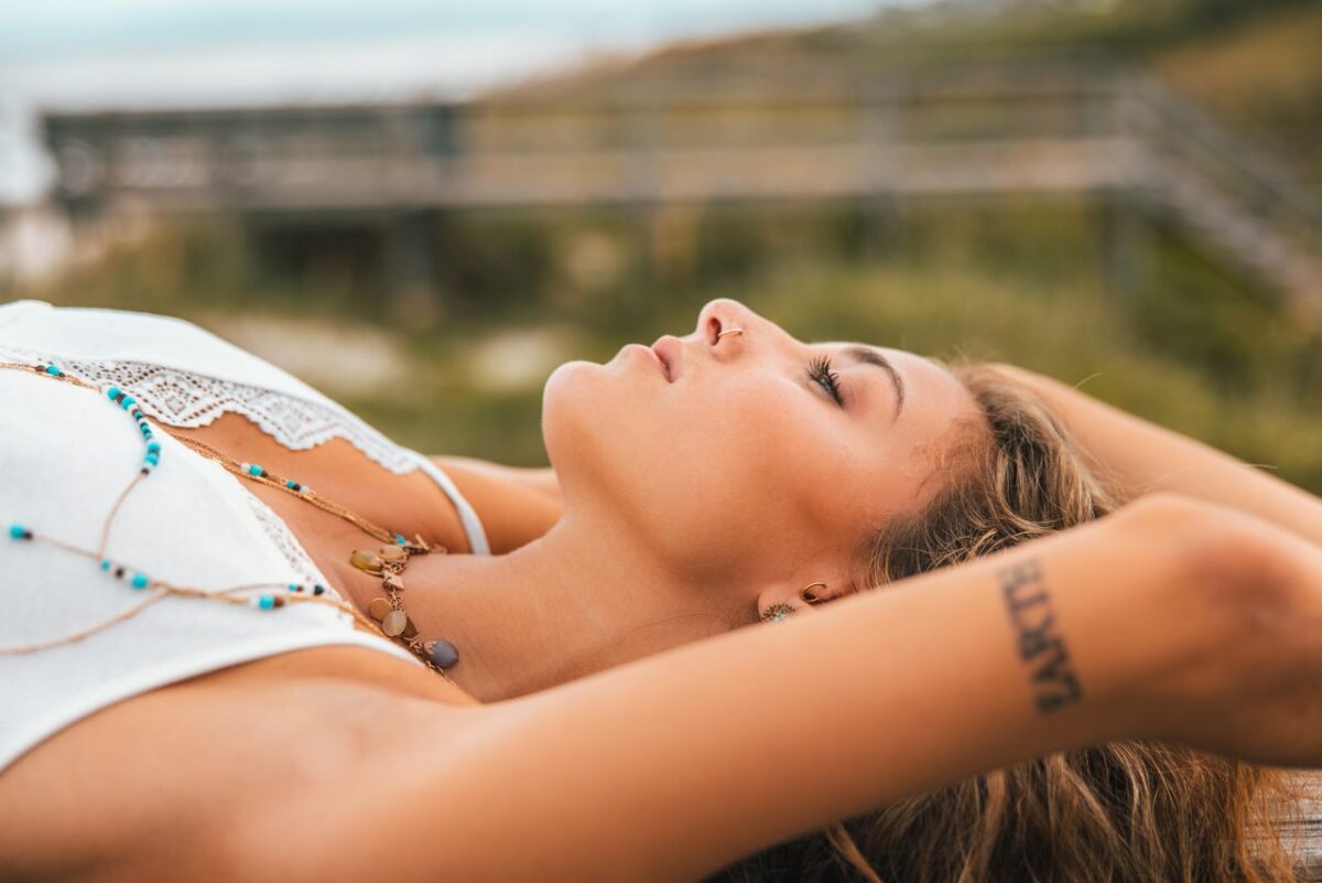 Young woman with tattoo lying outdoors, arms raised, in a peaceful pose.