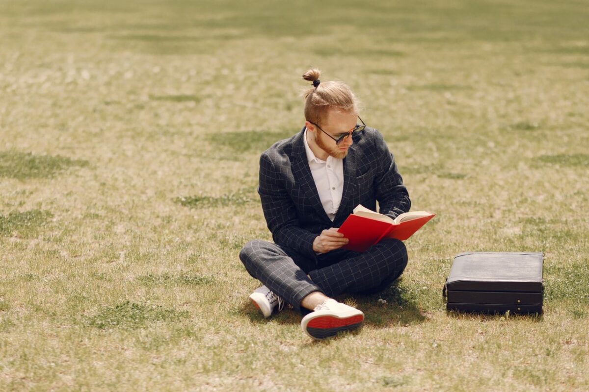 Young man in a suit reads a red book while sitting on grass in a sunny park.